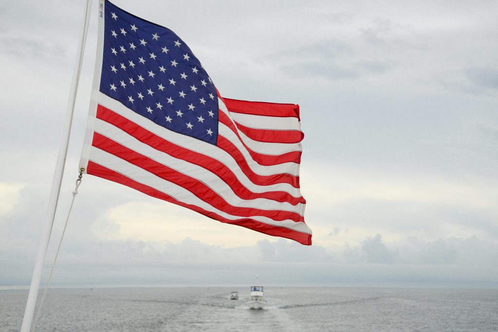 pexels-photo-11578702-11578702 A vibrant American flag flutters on a boat with a vast ocean and cloudy sky backdrop.